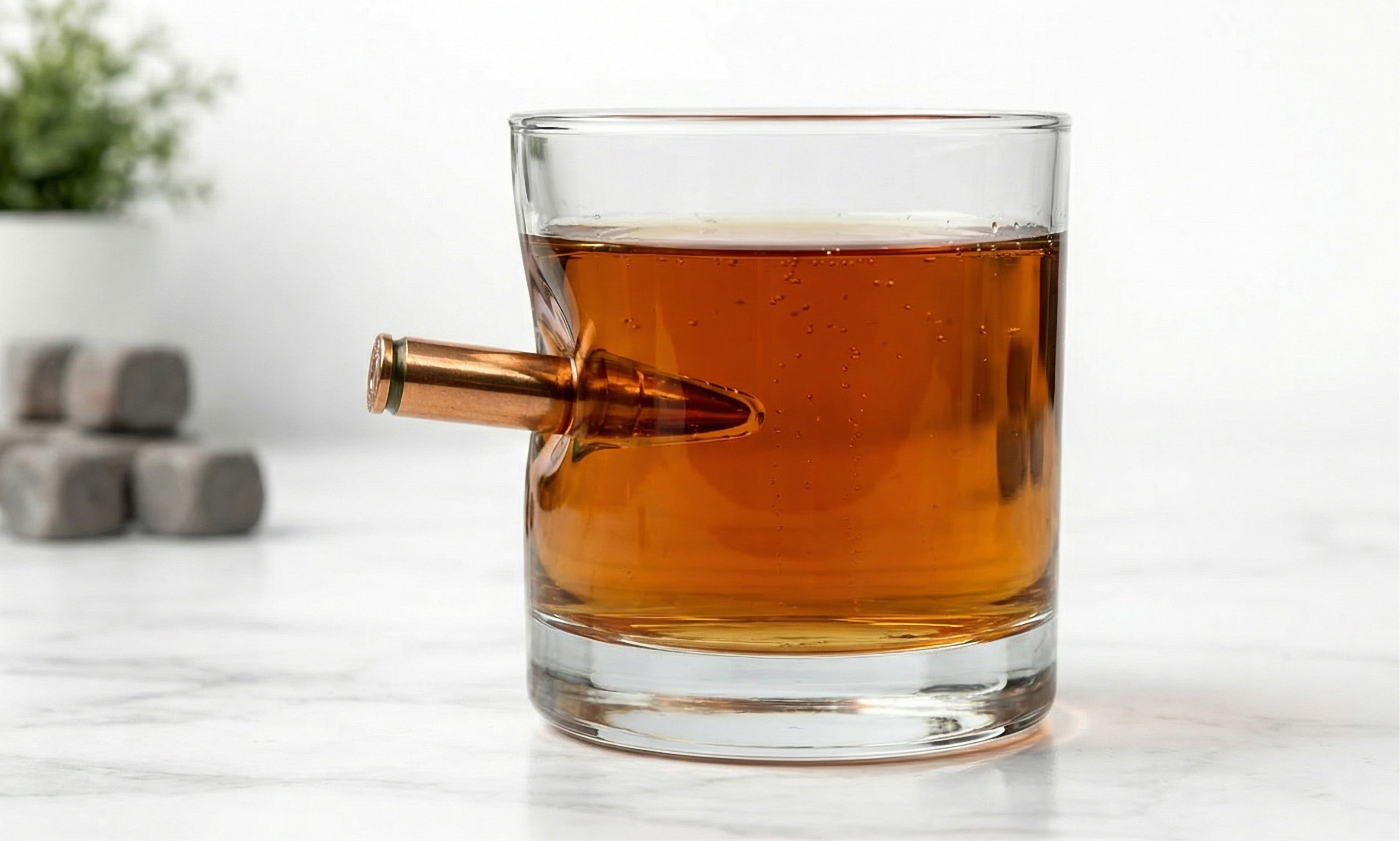 Ein elegantes Whiskeyglas gefüllt mit bernsteinfarbenem Whiskey, in dessen Seite eine kupferfarbene Patrone steckt. Im Hintergrund liegen graue Whiskey-Steine auf hellem Marmor.